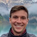 Brett, who is white and with short brown hair, smiling in front of a lake and mountains.